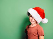 Positivo bambino che indossa il cappello rosso di Babbo Natale sorridente ampiamente contro lo sfondo verde e guardando la fotocamera — Foto stock