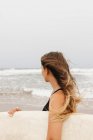 Side view of unrecognizable young sportswoman in swimwear with surfboard looking away on sandy coast against stormy ocean — Stock Photo