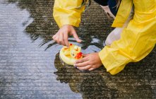 From above crop kid in raincoat playing with plastic ducks reflecting in rippled puddle in rainy weather — Stock Photo