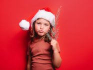 Cute little girl pouting lips in casual clothes and Santa hat holding fir tree twig and looking at camera against red background — Stock Photo