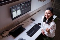 From above smiling ethnic female broker typing on keyboard against monitor with graphics and showing like gesture while looking at camera at home — Stock Photo