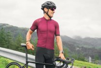 Vista lateral do desportista adulto alegre em óculos de sol de ciclismo e capacete sentado na bicicleta de estrada na estrada rural à luz do dia — Fotografia de Stock