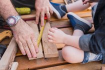 Crop unrecognizable dad measuring wooden piece with tape against child with pencil sitting on boardwalk — Stock Photo