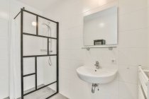 Мінімалістичний дизайн білої ванної кімнати з раковиною під дзеркалом, що звисає на плитці біля скляної душової кабіни в квартирі — стокове фото