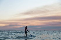 Вид ззаду на невизначений чоловічий серфер у гідрокостюмі та капелюсі на дошці для весла, що серфінгу на березі моря під час заходу сонця — стокове фото