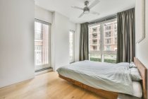 Bequemes Holzbett mit bequemer Decke und Kissen in der Nähe eines großen Fensters mit Vorhängen im Schlafzimmer — Stockfoto