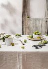 Still life composition of green fresh plums arranged with tableware on table covered with tablecloth — Stock Photo