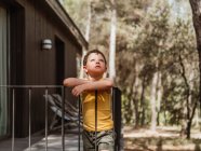 Seitenansicht eines kleinen Jungen, der im Sommer auf der Veranda eines modernen Ferienhauses im Wald steht — Stockfoto