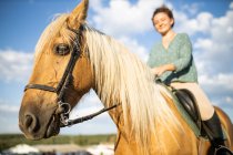 From below side view of female riding stallion with smooth brown coat on rough land against mount in countryside — Stock Photo