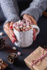 De arriba de la cosecha hembra sosteniendo taza de bebida caliente con malvaviscos entre los conos de regalos de Navidad y palitos de canela - foto de stock
