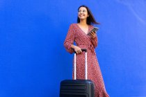 Positive Frau im langen roten Kleid, die mit Gepäck unterwegs ist, während sie tagsüber mit dem Smartphone auf der Straße gegen eine blaue Wand surft — Stockfoto