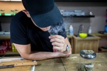 Anonyme adulte mâle en casquette fumant du cannabis joint dans l'espace de travail sur fond flou — Photo de stock