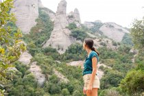 Vista lateral del viajero femenino con las manos en las caderas contemplando Montserrat con árboles mientras mira hacia otro lado durante la excursión en España - foto de stock