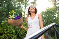 From below content young female in eyeglasses looking away standing with blossoming flower bouquet on urban stairs — Stock Photo
