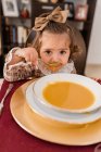 Очаровательный ребенок с луком на каштановых волосах и ложкой, смотрящий в камеру на тарелку супа из пюре для сквоша в доме — стоковое фото