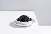 Estúdio minimalista com espaguete de tinta de lula preta em tigela de cerâmica completa na mesa branca — Fotografia de Stock