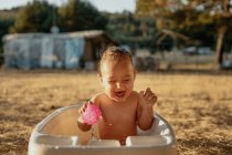 Happy toddler child with closed eyes and toy sitting in plastic bath while playing with water in countryside — Stock Photo