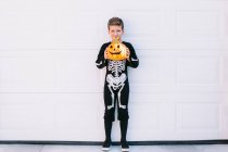 Full body of smiling preteen boy wearing black Halloween costume with skeleton print standing near carved Jack O Lantern pumpkin against white wall — Stock Photo