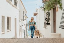 Verträumte Frau mit Korbtasche und Sonnenbrille in der Hand spaziert auf gepflasterter Straße zwischen weißen Häusern in der Küstenstadt — Stockfoto