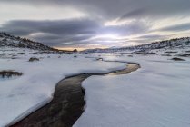 Пейзаж сніжного схилу пагорба в високогір'ї під хмарним небом вдень і річкою крижаної води — стокове фото