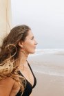Vue latérale d'une jeune athlète réfléchie en maillot de bain avec des cheveux volants et une planche de surf regardant loin sur la plage de l'océan — Photo de stock