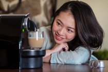 Crop young ethnic female leaning on hands against pod coffee maker pouring hot beverage with foam into glass in house kitchen — Stock Photo