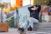 Full body of positive young female wearing casual black hoodie and light blue jeans with slits and roller blades sitting on back of wooden bench in skate park — Stock Photo