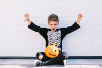 Corpo inteiro de garoto animado em traje de esqueleto com maquiagem e abóbora de Halloween esculpida levantando braços e gritando com rosto assustador enquanto sentado perto da parede branca — Fotografia de Stock