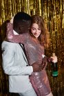 Freundliche junge Frau mit Flasche Champagner umarmt Afroamerikaner, während er sie umarmt und ihr während der Party Dinge ins Ohr sagt — Stockfoto