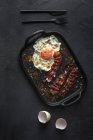 Vista dall'alto dell'uovo soleggiato con fette di pancetta fritte e condimenti su vassoio contro posate su sfondo scuro — Foto stock