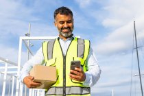 From below pleasant bearded ethnic male worker in uniform carrying parcel while using mobile phone at work — Stock Photo