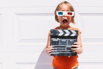 Cute astonished girl in casual colorful clothes holding clapperboard while on three dimensional glasses standing on white wall background — Stock Photo