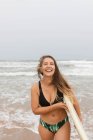 Allegro giovane sportiva in costume da bagno con tavola da surf guardando la fotocamera sulla costa sabbiosa contro l'oceano tempestoso — Foto stock