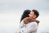 Vista lateral do homem sorridente abraçando namorada indiana em pé no campo sob céu nublado — Fotografia de Stock
