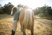 Seitenansicht einer Hündin mittleren Alters mit lockigem Haar, die einen Sattel auf den Rücken der Stute legt, auf einem Bauernhof im Sonnenlicht — Stockfoto