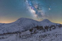 Paysage à couper le souffle de pente de colline couverte de neige et d'arbres contre de hautes montagnes rocheuses sous un ciel étoilé nocturne avec la Voie lactée — Photo de stock