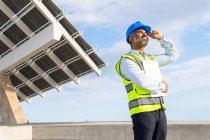 Low angle of mature ethnic male inspector in vest and hardhat with clipboard looking away while standing near solar power station — Stock Photo