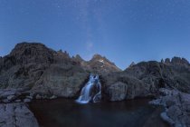 Захватывающие пейзажи грубых скалистых образований с водопадом, струящимся к озеру под безоблачным звездным небом ночью — стоковое фото