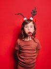 Joyful little girl in casual clothes and festive deer headband blowing party whistle and looking at camera during Christmas celebration against red background — Stock Photo