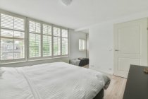 Удобная мягкая кровать с белым одеялом, расположенная рядом с большими окнами в современной спальне в квартире — стоковое фото
