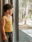 Heiteres kleines Kind steht am Fenster in Hütte und schaut in Gedanken weg — Stockfoto