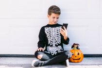 Full body of cheerful boy in black skeleton costume with painted face and carved Halloween pumpkin using on mobile phone while sitting near white wall on street — Stock Photo