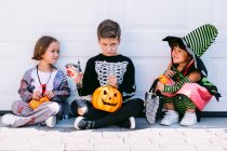 Full body of group of little kids dressed in various Halloween costumes with carved Jack O Lantern sitting near white wall on street — Stock Photo