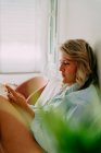 Side view of adult female in shirt surfing internet on mobile phone while sitting in light room — Stock Photo