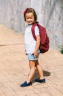 Seitenansicht eines Schulkindes mit Rucksack auf dem Bürgersteig, das im Sonnenlicht in die Kamera blickt — Stockfoto