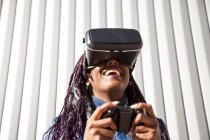 Jeune femme afro-américaine excitée dans un casque VR en utilisant un contrôleur tout en divertissant et en jouant à un jeu virtuel contre un mur rayé gris — Photo de stock