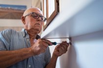 Maschio anziano attento in occhiali con cacciavite manuale avvitando mensola a parete in camera di casa — Foto stock