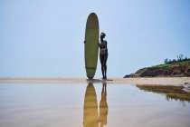 Vue de face de l'athlète afro-américaine avec planche de surf admirant l'océan depuis le rivage sablonneux sous un ciel bleu nuageux — Photo de stock