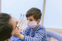 Neugieriger Junge in medizinischer Maske schlüpft in die Rolle des Zahnarztes und checkt Zähne mit Zahnspiegel im Krankenhaus — Stockfoto