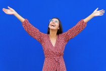 Mujer joven positiva con el pelo castaño en vestido rojo de pie con los brazos levantados y mirando hacia arriba contra el fondo azul en el día - foto de stock